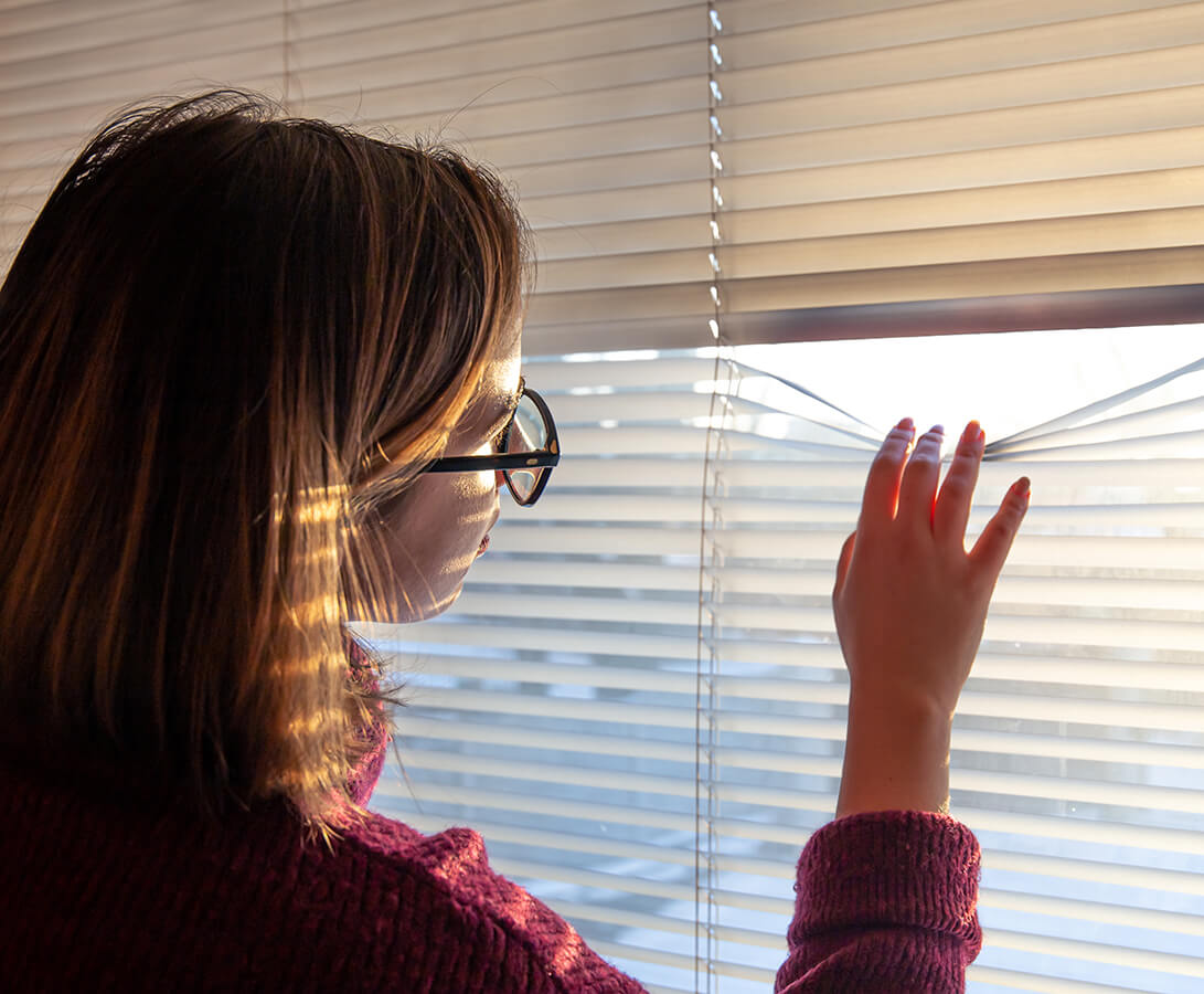 A woman looking out through her shut blinds.