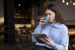 A woman looking at a tablet while drinking a hot beverage.